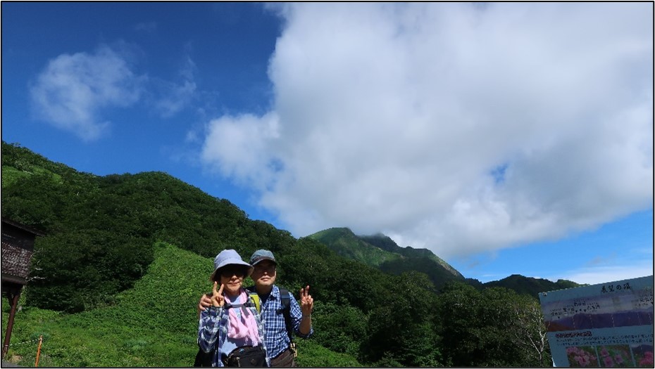 緑豊かな山々を背景に二人がピースサインをしている写真