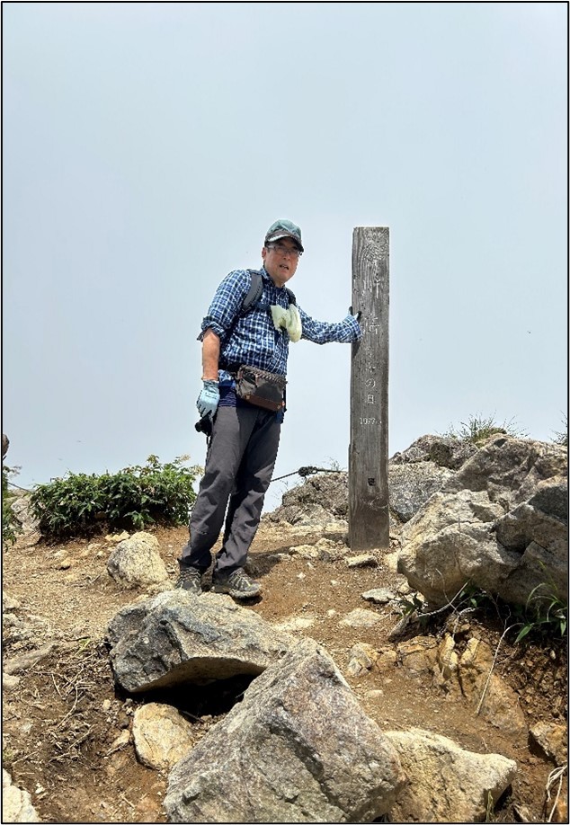 山頂で木製の標柱に手をかけて立つ登山者の写真